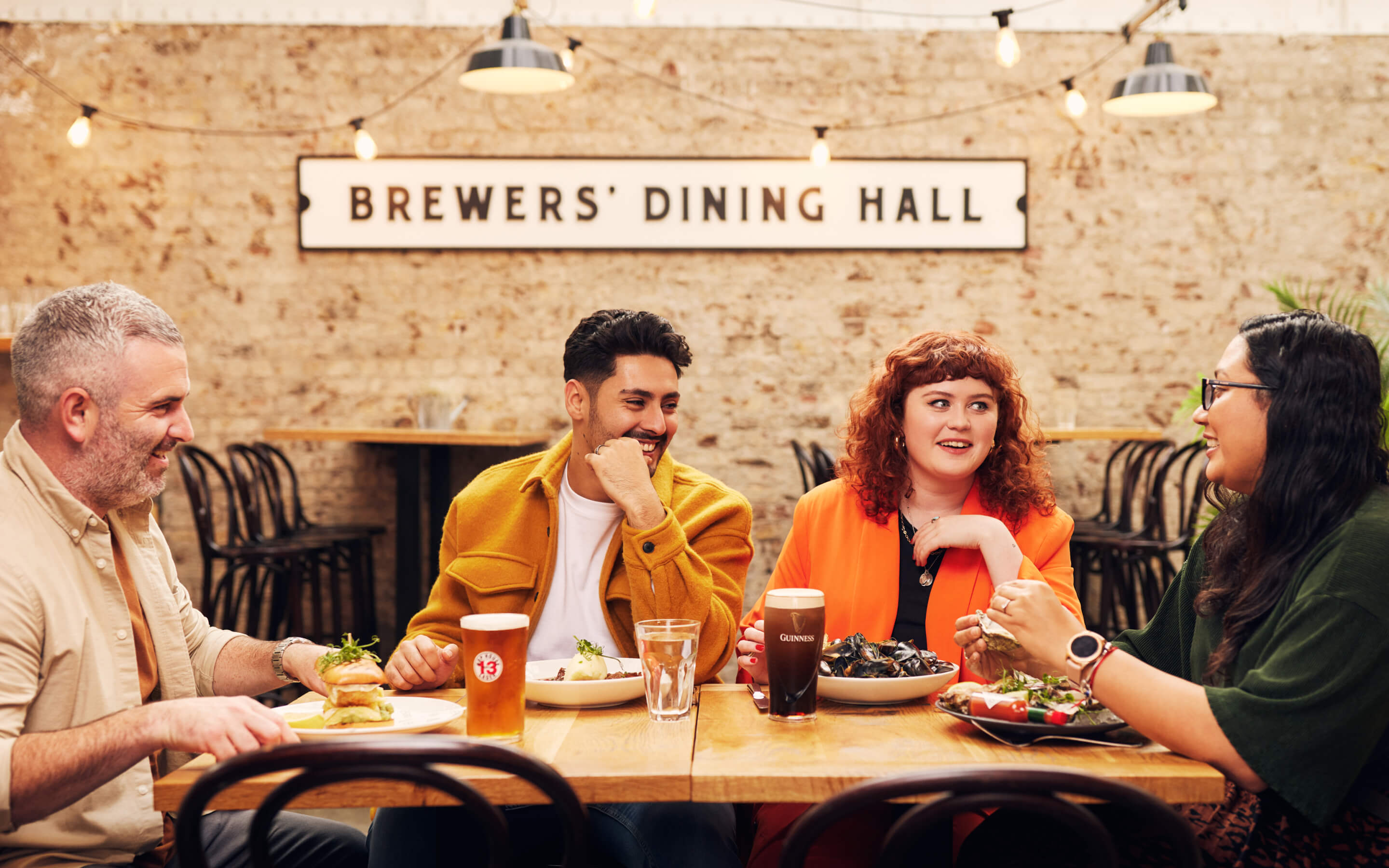 Group of friends laughing and enjoying their meal at the Brewers’ Dining Hall, one with the pairing of mussels & Guinness and another with hake & Hop House 13.